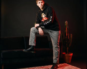 Peut inclure: Un sweat à capuche noir avec une illustration d'un personnage d'anime portant un chapeau de paille sur la poitrine et une série de petites icônes de crânes sur la manche gauche, associé à un jean en denim gris et des bottes en cuir noir.