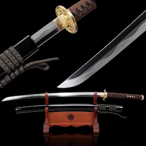 Puede incluir: Una espada katana con hoja de acero curva, empuñadura envuelta en cordón marrón y guarda circular dorada. La espada se exhibe sobre un soporte de madera junto a una vaina lacada en negro con detalles decorativos de cordón marrón.