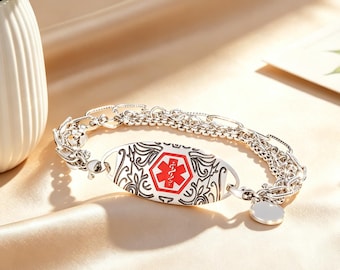 May include: A silver-toned medical alert bracelet featuring a central oval plaque with an engraved floral pattern and a red Star of Life symbol. The bracelet is constructed with a multi-strand chain design and includes a small, round, blank silver charm.