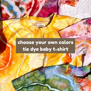 May include: Three short-sleeve baby t-shirts featuring tie-dye patterns in vibrant shades of pink, orange, yellow, green, and blue. The shirts have a round neckline and a small interior label reading earth to jude. Text overlay reads: choose your own colors tie dye baby t-shirt.