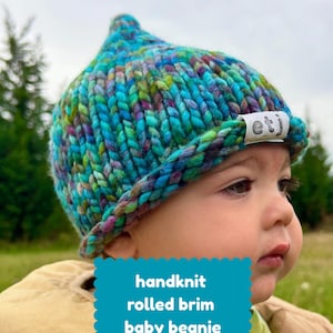 May include: A hand-knit baby beanie with a rolled brim, crafted from chunky yarn in a variegated color palette of teal, blue, purple, and green. A small white fabric tag on the brim features the black text etj. The beanie has a pointed top.