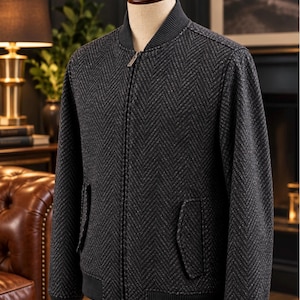 May include: A dark gray bomber jacket featuring a textured herringbone pattern, a ribbed collar, ribbed cuffs, and a full-length front zipper. The jacket includes two side pockets with flaps and is displayed on a mannequin torso.