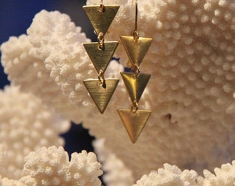May include: A pair of gold triangle earrings with a cascading design. The earrings are hanging from a white coral background.
