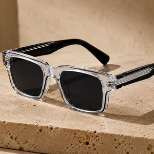 May include: Sunglasses featuring a clear, transparent rectangular frame with dark, solid black lenses. The temples are black and transition into a ribbed, clear plastic detail near the hinges, creating a distinct contrast in color and texture.