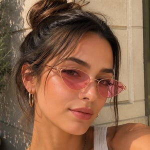 May include: Cat-eye sunglasses with thin gold-tone metal frames and translucent pink lenses. The eyewear features a sharp, angular silhouette that tapers at the outer edges, paired with a simple gold-tone bridge and slender temples.