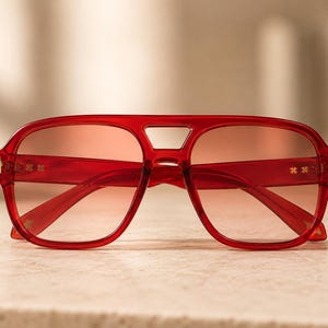 Puede incluir: Gafas de sol tipo aviador con montura roja translúcida y lentes rojas degradadas. La montura presenta un diseño de doble puente y pequeños detalles metálicos en tono dorado en las patillas y bisagras, con una forma clásica de piloto.