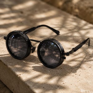 May include: Black round sunglasses with a steampunk design. The sunglasses have dark lenses and a matte black frame. The frames have a unique, layered design around the lenses and the arms are hinged. The sunglasses are resting on a light-coloured surface.
