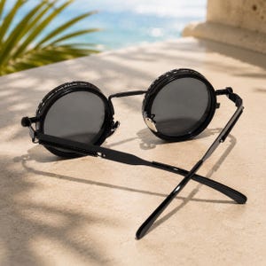 May include: Black round sunglasses with dark grey lenses. The sunglasses have a unique design with a thin black frame and a spring hinge. The lenses are set in a circular frame with decorative detailing. The sunglasses are resting on a light-coloured surface.
