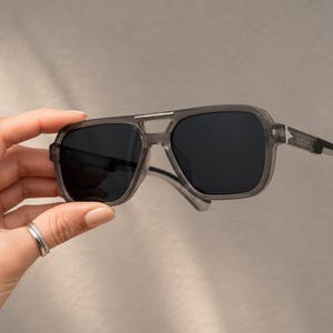 Peut inclure: Lunettes de soleil grises avec des verres foncés. Les lunettes ont une forme carrée avec un double pont et des branches épaisses. La monture est de couleur grise translucide.