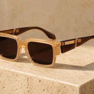 Peut inclure: Lunettes de soleil avec une monture carrée beige translucide et des verres marron foncé. Les branches présentent une finition marron foncé avec un accent métallique doré rectangulaire près de la charnière. Le design inclut un pont épais.