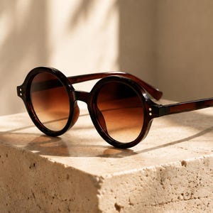 Peut inclure: Lunettes de soleil rondes avec une monture en écaille de tortue marron foncé et des verres dégradés passant d'un marron foncé en haut à une teinte ambrée plus claire en bas. La monture présente de petits rivets métalliques argentés aux charnières.