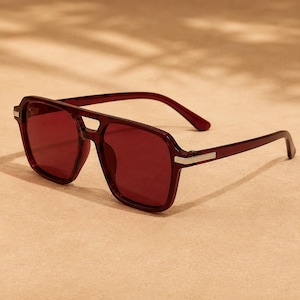 Peut inclure: Lunettes de soleil bordeaux avec une monture carrée et un double pont. Les verres et les montures sont d'un rouge profond assorti, avec des accents argentés sur les branches. Les lunettes sont posées sur un fond neutre.