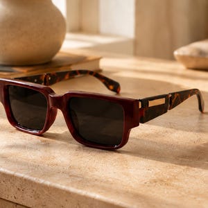 Peut inclure: Lunettes de soleil rectangulaires dotées d'une monture bordeaux foncé et de verres teintés sombres. Les branches présentent un motif écaille de tortue dans des tons marron et noir, rehaussé par un petit détail métallique doré au niveau de la charnière.