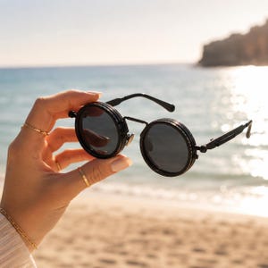 May include: Black round sunglasses with dark lenses. The sunglasses have a unique double-rimmed frame and a sleek, modern aesthetic. The sunglasses are held up against a beach background.