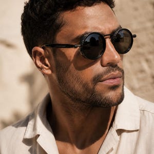 May include: Round black sunglasses with a dark frame. The sunglasses have a unique design with side shields. The person in the image is wearing a light beige button-down shirt.