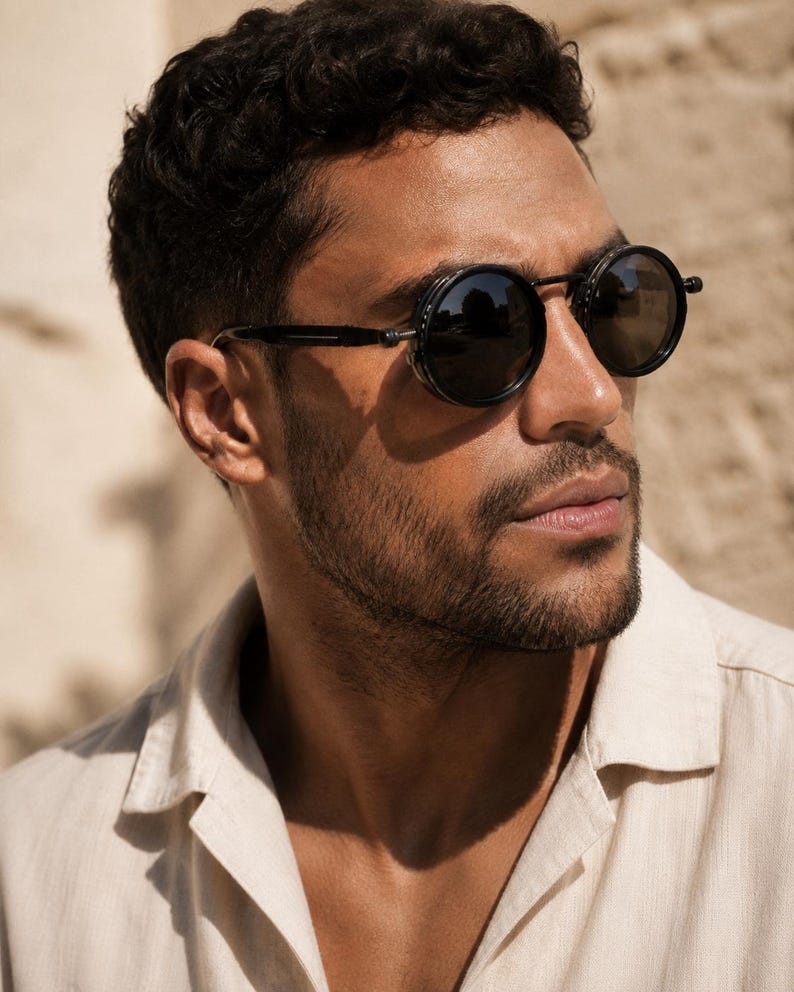 May include: Round black sunglasses with a dark frame. The sunglasses have a unique design with side shields. The person in the image is wearing a light beige button-down shirt.