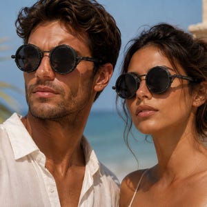 May include: A man and a woman wearing round, black sunglasses with dark frames. The sunglasses have a vintage or steampunk aesthetic. The man is wearing a white button-down shirt, and the woman is wearing a light-coloured top.