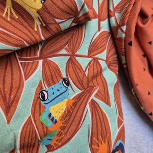 Puede incluir: Una tela con un patrón repetitivo de ranas y hojas. El diseño incluye ranas amarillas y azules con extremidades naranjas posicionadas entre follaje marrón sobre un fondo verde menta claro. El tejido presenta una textura suave y plegada.