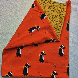 Peut inclure: Un tour de cou en tissu plié présentant un extérieur orange avec un motif répétitif de blaireaux noirs et blancs portant des écharpes jaunes et de petits champignons jaunes. La doublure intérieure est un tissu jaune moutarde à pois noirs.