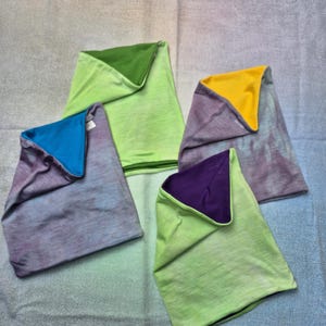 Op de afbeelding: Vier omkeerbare nekwarmers met een tie-dye patroon aan de ene kant en een effen kleur aan de andere kant. De kleuren omvatten limoengroen, felblauw, geel en dieppaars, waarbij elk stuk een unieke combinatie van deze tinten laat zien.