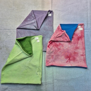 Op de afbeelding: Drie gevouwen stoffen nekwarmers. Eén is lichtpaars met een effen paarse binnenkant. Eén is lichtgroen met een effen groene binnenkant. Eén heeft een roze en wit tie-dye patroon met een effen blauwe binnenkant. Elk heeft een klein wit label.