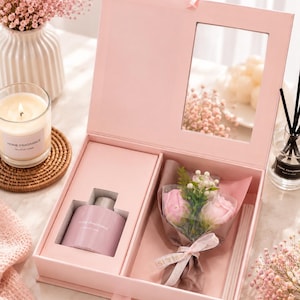 May include: A light pink rectangular gift box with a hinged lid and a clear window. Inside, a mauve fragrance bottle sits in a custom cutout next to a small bouquet of two pink artificial roses wrapped in netting and tied with a pink ribbon.