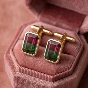 Puede incluir: Un par de gemelos dorados con piedras preciosas rectangulares de corte esmeralda que presentan un degradado de color desde el magenta intenso en la parte superior hasta un verde bosque vibrante en la base, engastados en marcos de metal dorado.