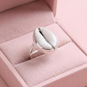 Könnte beinhalten: Ein silberner Ring mit einer großen, weißen Kaurischnecke. Die Schnecke ist der Mittelpunkt, eingebettet in ein poliertes Silberband. Der Ring wird in einer rosa Samt-Schmuckschatulle präsentiert, die das Design und die Farbe des Rings hervorhebt.