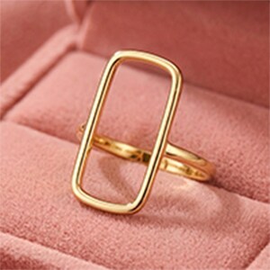 May include: A gold-toned ring featuring a large, vertical, open rectangular frame as the centrepiece. The band is thin and circular, connecting to the base of the smooth, polished rectangular wire structure that stands upright on the finger.