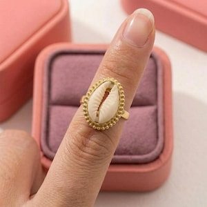 Könnte beinhalten: Ein goldfarbener Ring mit einer mittig platzierten, ovalen weißen Kaurimuschel. Die Muschel ist von einem zarten, perlenartigen goldfarbenen Metallrand umgeben. Der Ring ist schmal und metallisch und weist eine polierte Oberfläche auf, die die natürliche Textur der Muschel unterstreicht.