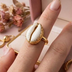 Könnte beinhalten: Ein goldfarbener Ring mit einer zentralen weißen Kaurimuschel, die in eine texturierte, gehämmerte Metallfassung eingefasst ist. Die Ringschiene ist glatt und poliert und zeigt einen deutlichen Kontrast zwischen der glänzenden Muschel und dem Gold.