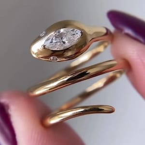 Peut inclure: Une bague en forme de serpent doré avec une pierre claire taille marquise sur la tête et deux petites pierres rondes claires en guise d'yeux. L'anneau s'enroule en spirale et présente une finition métallique polie sur toute la surface.