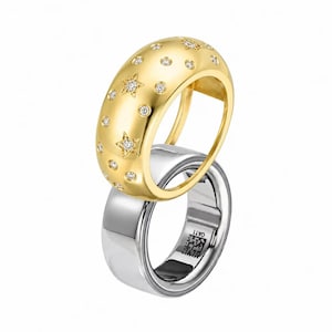 Peut inclure: Une bague dorée à surface bombée ornée d'étoiles et de cercles incrustés, chacun serti d'une petite pierre claire. Cette bague dorée est placée au-dessus d'un anneau argenté lisse et uni portant une petite gravure de code QR carré.