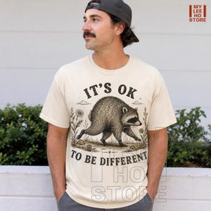 May include: A cream-colored short-sleeve t-shirt featuring a black ink-style illustration of a raccoon walking through a field of flowers. The text above the raccoon reads IT'S OK and the text below reads TO BE DIFFERENT in a bold, dark font.