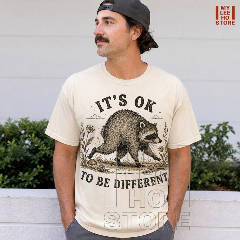 May include: A cream-colored short-sleeve t-shirt featuring a black ink-style illustration of a raccoon walking through a field of flowers. The text above the raccoon reads IT'S OK and the text below reads TO BE DIFFERENT in a bold, dark font.