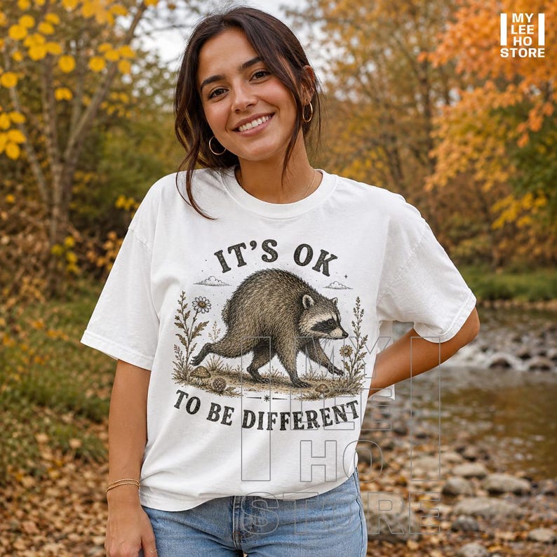 May include: A white short-sleeve t-shirt featuring a graphic illustration of a raccoon walking among wildflowers. The text "IT'S OK TO BE DIFFERENT" is printed in black capital letters above and below the raccoon. The design is centered on the chest.
