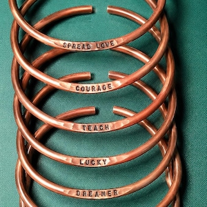 May include: A stack of six open-ended copper cuff bracelets with a hammered texture. Each bracelet is stamped with a unique word: SPREAD LOVE, COURAGE, TEACH, LUCKY, DREAMER, PRAY, and BE STRONG. The metal has a warm, metallic reddish-brown finish.