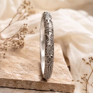 May include: A silver-toned metal bangle bracelet featuring an intricate, embossed filigree pattern across its outer surface. The bracelet has a rounded, polished profile with a visible hinge mechanism near the top, showcasing detailed metalwork.