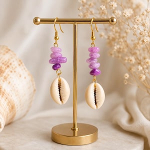 May include: A pair of dangle earrings featuring gold-toned hooks, each with a vertical stack of three smooth, graduated purple beads in varying shades of violet and lavender, finished with a single cream-colored cowrie shell at the bottom.