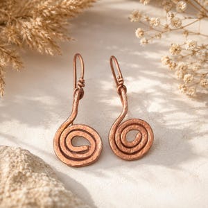 Könnte beinhalten: Ein Paar Ohrringe aus Kupfer mit einem gehämmerten, spiralförmigen Design. Jeder Ohrring hängt an einem einfachen Haken, wobei die Metalloberfläche ein strukturiertes, rustikales Finish aufweist. Die Spirale bildet eine kreisförmige Form.