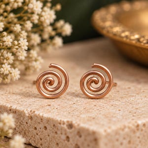 May include: A pair of rose gold stud earrings featuring a simple, continuous spiral design. The earrings are crafted from smooth, polished metal wire that curves inward to form a circular coil shape, creating a clean and symmetrical appearance.