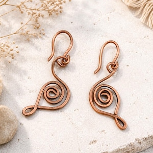 Puede incluir: Un par de pendientes de alambre de cobre con un diseño de espiral formado a mano. Cada pendiente consta de una sola pieza continua de alambre de metal moldeada en una bobina circular que termina en un gancho curvado para la oreja.