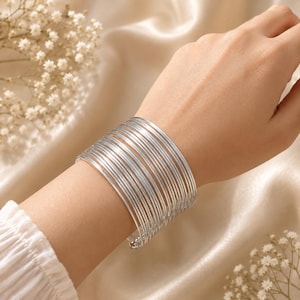 Op de afbeelding: Een brede armband bestaande uit meerdere dunne, gepolijste zilverkleurige draadringen die in een parallelle, gestapelde formatie zijn gerangschikt. De armband heeft een gladde, reflecterende metalen afwerking en een doorlopend, cirkelvormig ontwerp.