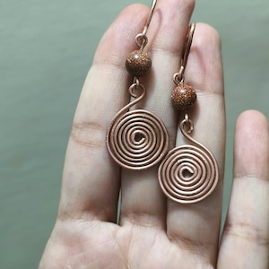 Puede incluir: Un par de pendientes colgantes con alambre de color cobre formado en espirales circulares planas. Cada pendiente está acentuado con una cuenta redonda de piedra de sol marrón brillante colocada sobre la espiral, suspendida de un gancho de metal.