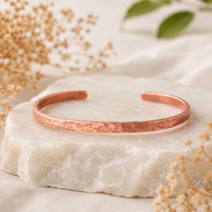 Pode incluir: Uma pulseira de cobre tipo bracelete com um acabamento martelado e texturizado. A pulseira é uma banda circular aberta com um tom castanho-avermelhado metálico e uma largura consistente em todo o design, exibindo uma superfície rústica.