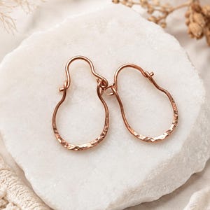 May include: A pair of rose gold hoop earrings with a distinct pear-shaped silhouette and a hammered, textured finish. The earrings feature a simple hook closure mechanism at the top, showcasing a metallic lustre and a reflective surface.