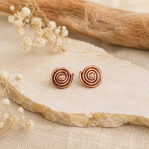 Puede incluir: Un par de pendientes de botón de color cobre con un diseño de espiral circular. Los pendientes están construidos con un solo alambre continuo enrollado en un patrón concéntrico apretado, creando una forma redondeada con acabado metálico.