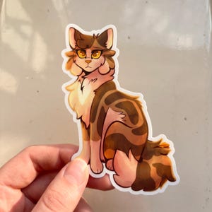 May include: A die-cut sticker featuring a digital illustration of a brown and cream-colored tabby cat with large, expressive amber eyes. The cat is depicted in a seated position with fluffy fur, dark brown patches, and a white chest, outlined in white.