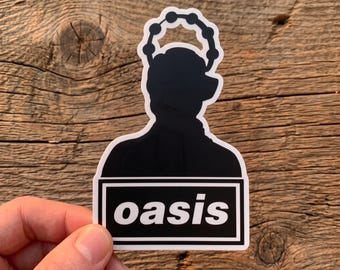 Könnte beinhalten: Ein gestanzter Aufkleber mit der schwarzen Silhouette einer Person, die einen Tamburin-Heiligenschein über dem Kopf trägt. Unter der Figur befindet sich ein schwarzes Rechteck mit weißem Rand, das das Wort oasis in weißen Kleinbuchstaben enthält.