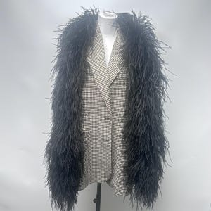 Op de afbeelding: Een blazer met een zwart-wit gingham ruitpatroon, geaccentueerd met lange, volumineuze zwarte struisvogelveren langs de revers en voorranden. Het kledingstuk heeft een reverskraag, een knoopsluiting aan de voorzijde en lange mouwen.